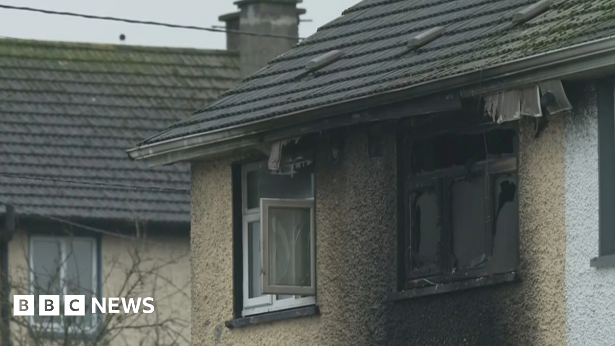 The camera is zoomed in on the top two windows of a light brown house. The window on the right id smashed and is black with burn marks. The area around the window is also stained black. The window to the right is open and relatively untouched by heat.