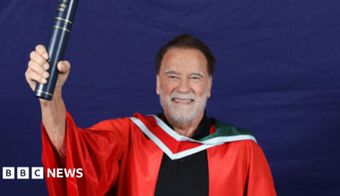 Arnold Schwarzenegger, a man with dark hair and a white beard, smiles while holding a navy and gold scroll holder in one hand.  He is wearing a red university gown with a white and red collar