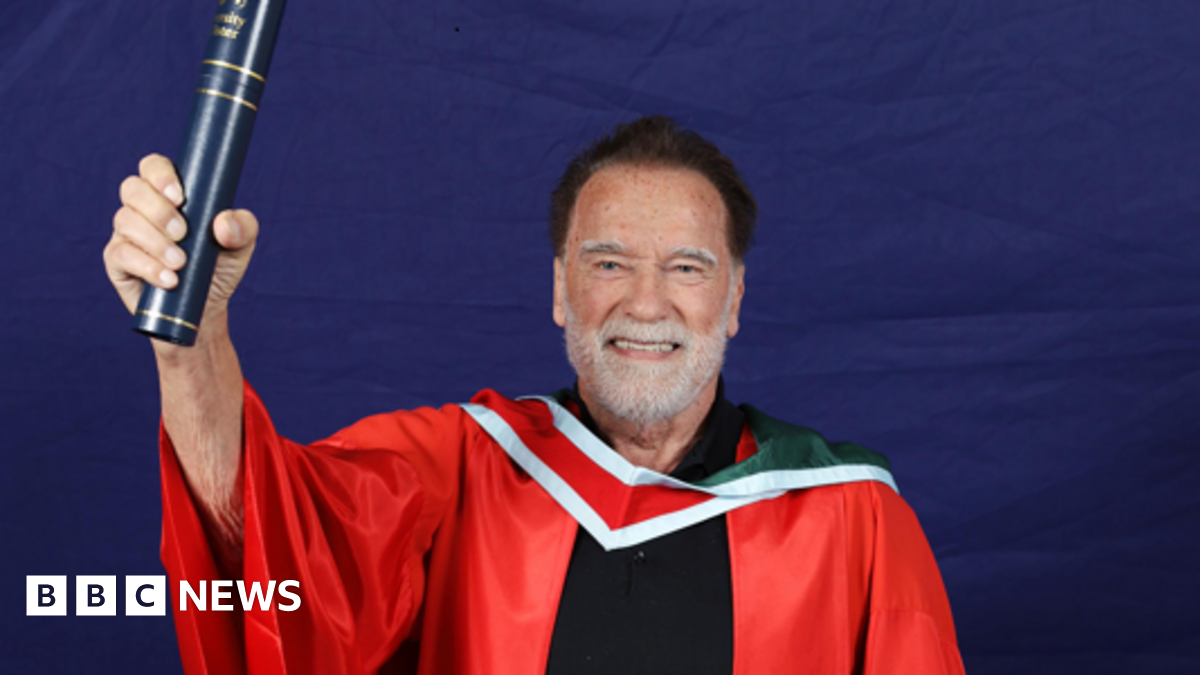 Arnold Schwarzenegger, a man with dark hair and a white beard, smiles while holding a navy and gold scroll holder in one hand.  He is wearing a red university gown with a white and red collar