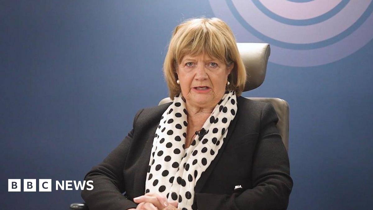 Baroness Heather Hallett sits in the inquiry chair wearing a black suit and spotty scarf, with the UK Covid-19 Inquiry logo on the wall behind her
