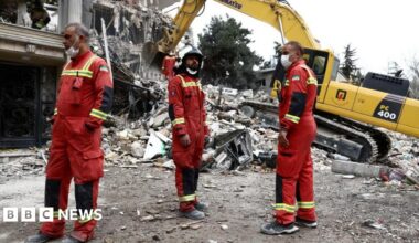 Three emergency workers in red uniforms stand near a yellow digger as it removes rubble