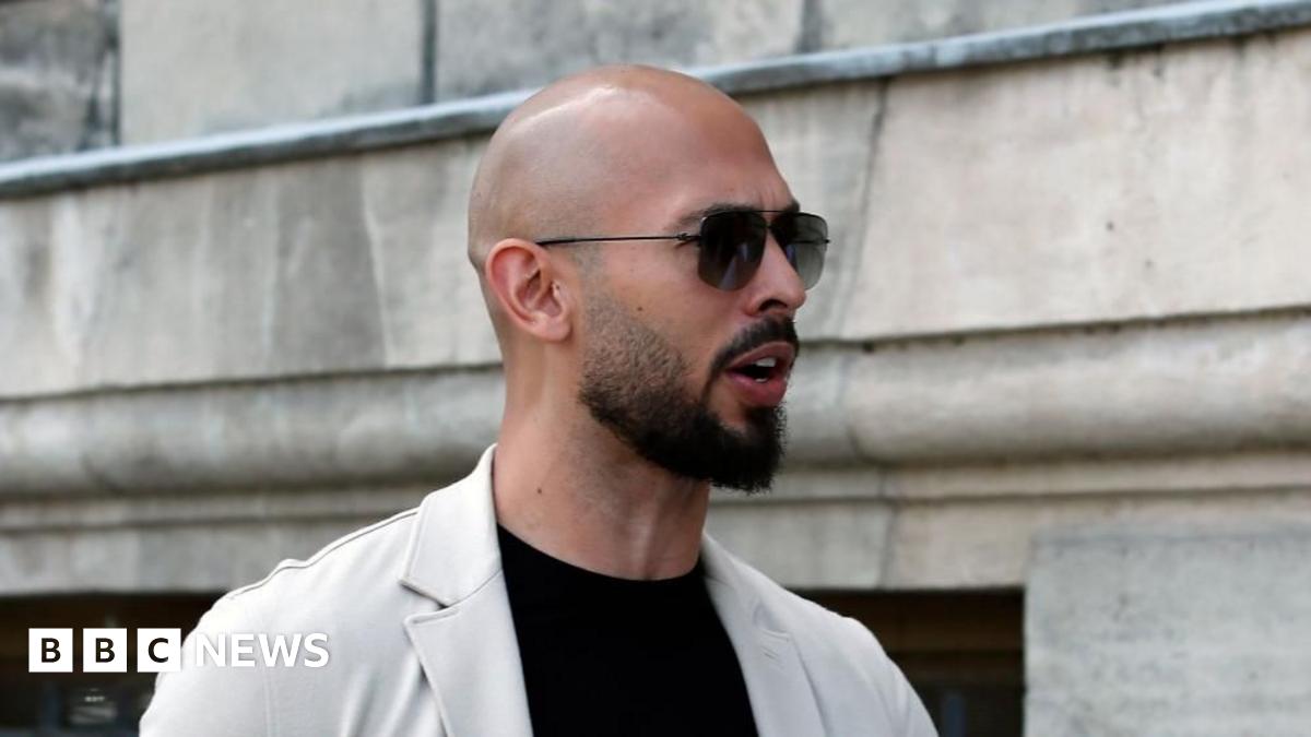 Andrew Tate pictured outside, wearing a black T-shirt with a beige jacket and a pair of sunglasses.