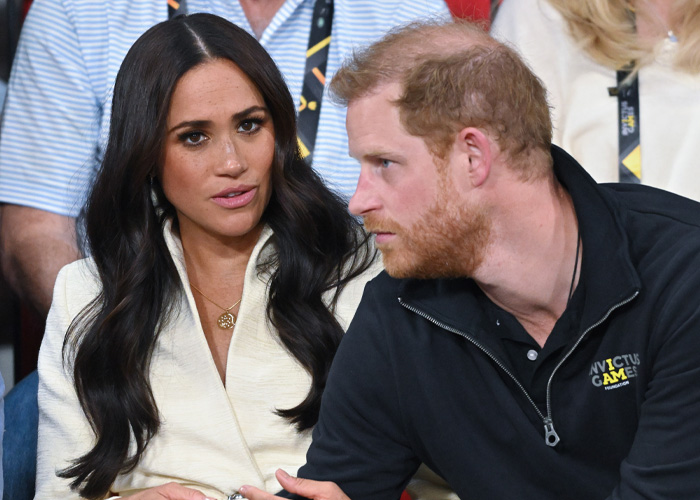 Harry and Meghan seated at an event, engaged in serious conversation amid a crowded background.