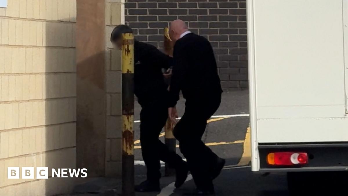 A man being led away from a van by another man wearing a black jumper and a shirt. He is walking into a brick building.