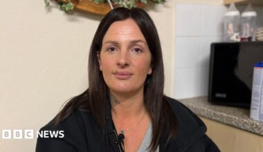 Krystal Ogier, a middle-aged woman, wearing a neutral expression. She is wearing a black hoodie with a grey T-shirt underneath. She has a tattoo on her neck and long, dark brown hair.