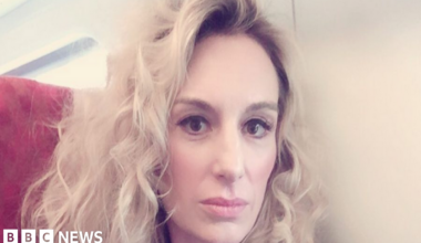 A woman with blonde, curly hair looks into camera. She is sat on a red chair with a white wall behind her. She has thick eyeliner on and is posing for the camera.