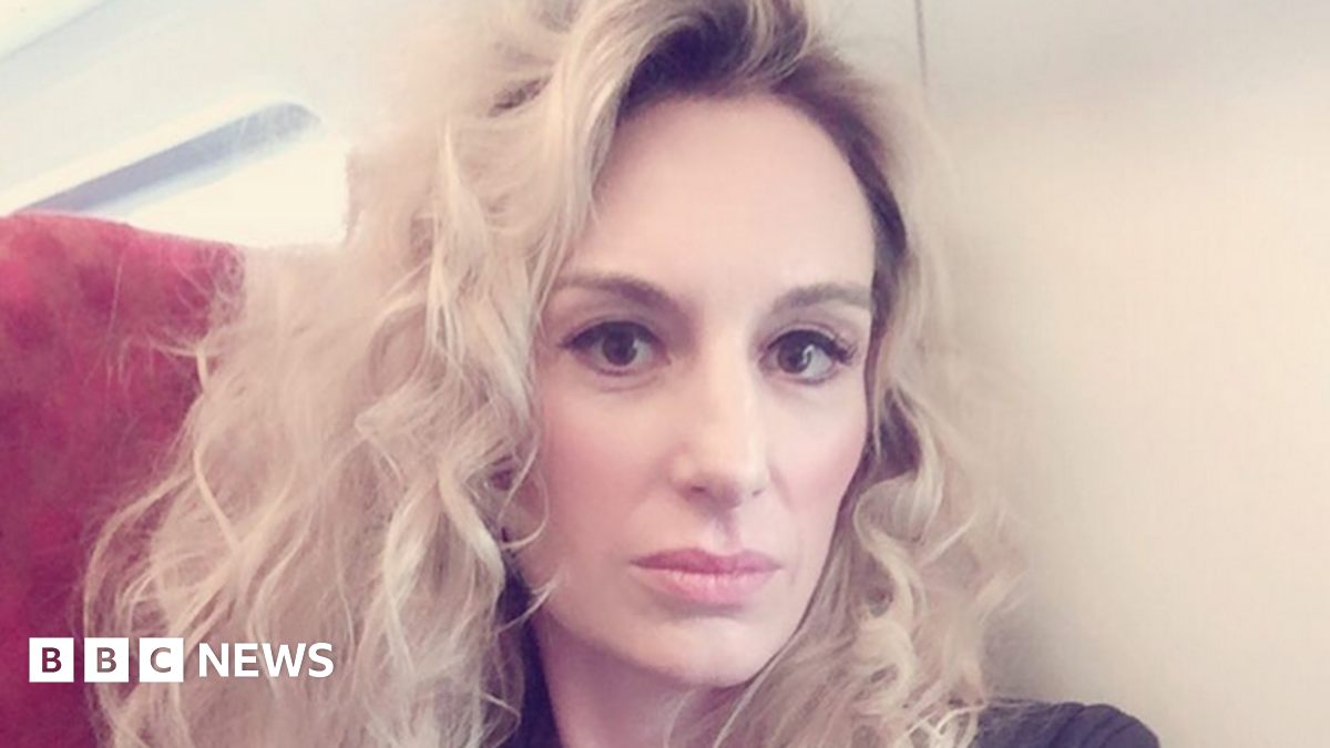 A woman with blonde, curly hair looks into camera. She is sat on a red chair with a white wall behind her. She has thick eyeliner on and is posing for the camera.