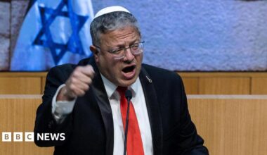 Itamar Ben-Gvir wearing a black suit, red tie and white kippah, talking on a podium in front of Israeli flags
