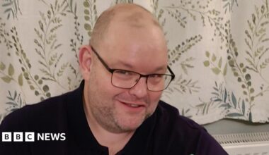A man, Gary Stanley, smiles into the camera. He is wearing black glasses and a black top. Behind him are white curtains with details of green leaves and branches.