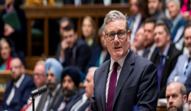 Keir Starmer standing in the House of Commons, while his MPs look on. He is wearing a dark suit and a purple tie.