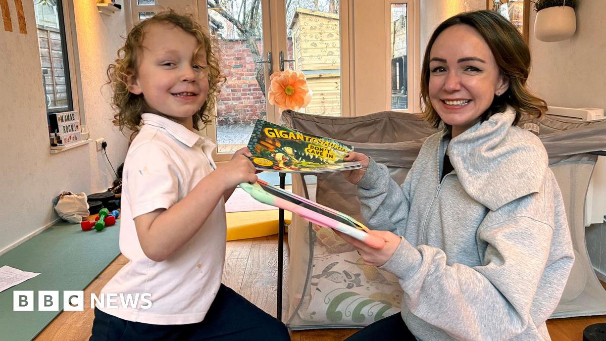 Romi, who has blond curly hair and is wearning a white t-shirt, is passing a book to his mum, Alexis who has short brown hair and is wearning a grey hoodie. She is passing a digital device with a colourful cover back to him and both are smiling.