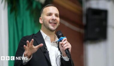 Zack Polanski, in white open necked shirt and dark jacket, addresses a Green Party rally, with microphone in hand
