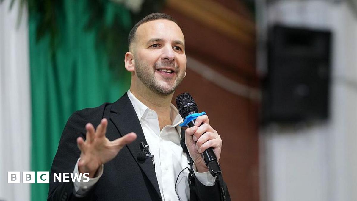 Zack Polanski, in white open necked shirt and dark jacket, addresses a Green Party rally, with microphone in hand