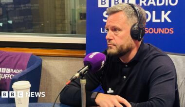 A man is sat in front of a purple microphone and wearing headphones inside a radio studio