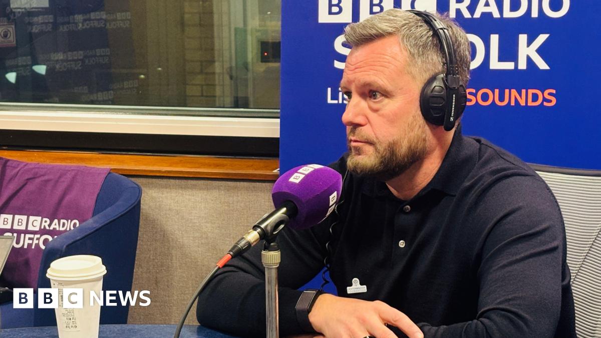 A man is sat in front of a purple microphone and wearing headphones inside a radio studio