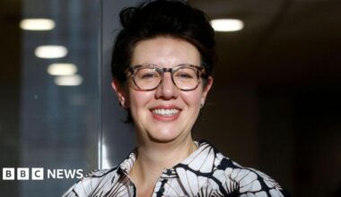 Claire Lynch smiling, wearing glasses and a black-and-white patterned shirt