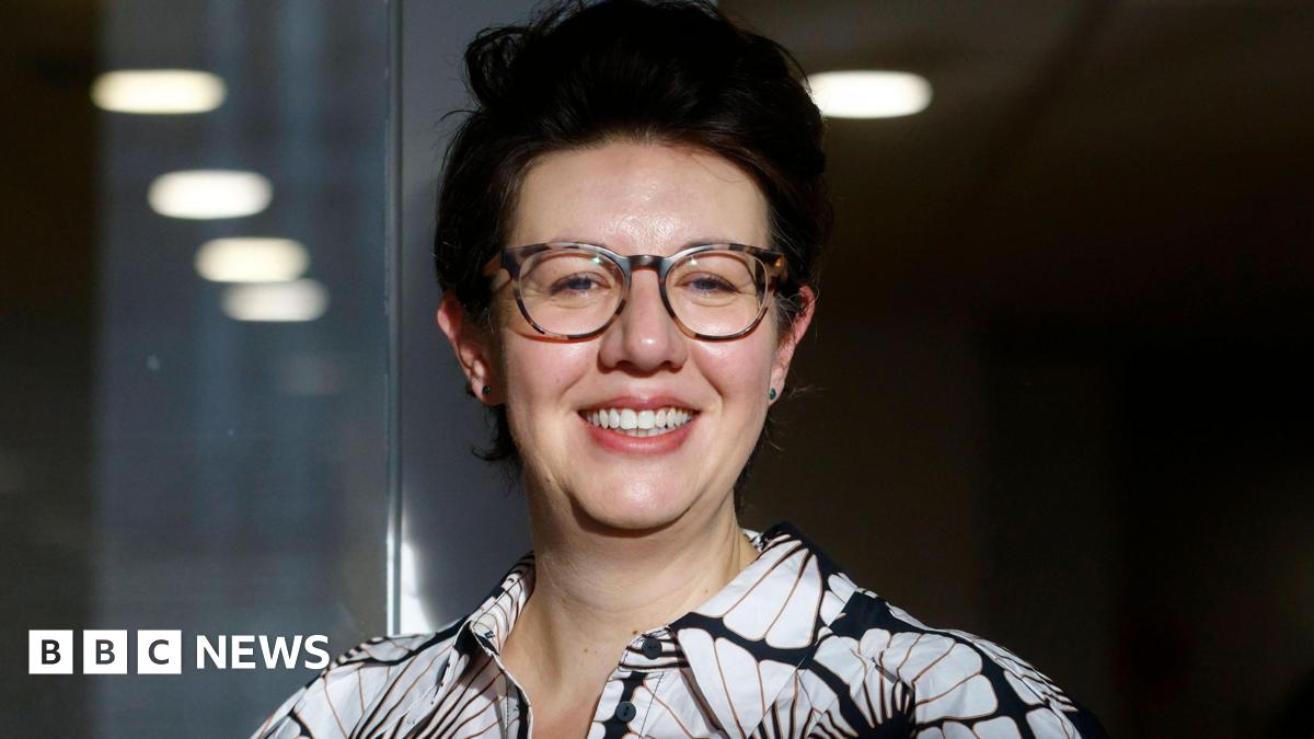 Claire Lynch smiling, wearing glasses and a black-and-white patterned shirt