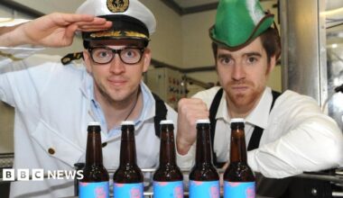 A man dressed as a sea captain next to a man wearing a green hat, posing for a photograph in front of bottles of beer