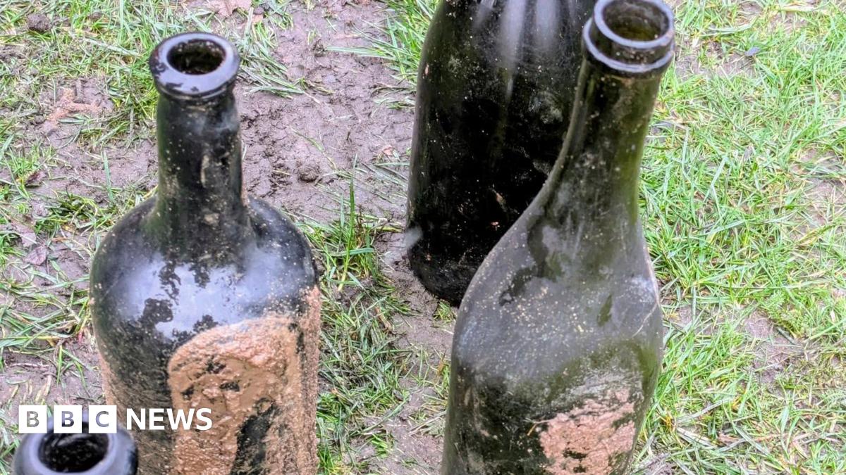 Four dark green gloss bottles, some encrusted with mud, standing on a muddy patch of grass
