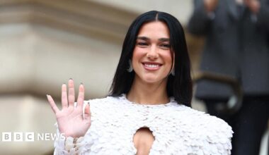 Dua Lipa wearing a feathered white outfit, waving at photographers and smiling.