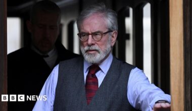 A man with glasses, white hair and a white beard pictured after passing through a doorway. He is wearing a shirt, tie and waistcoat. A man behind him can be seen cloaked in darkness.