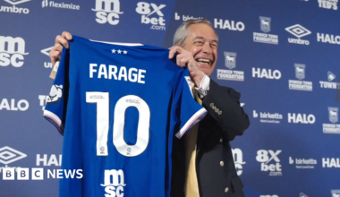 Nigel Farage poses with a blue Ipswich Town home shirt with 'FARAGE 10' on the back. He is standing in front of a blue advertising wall with various sponsors' logos on it. He is looking away from the camera and laughing.