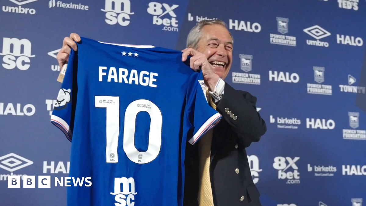Nigel Farage poses with a blue Ipswich Town home shirt with 'FARAGE 10' on the back. He is standing in front of a blue advertising wall with various sponsors' logos on it. He is looking away from the camera and laughing.