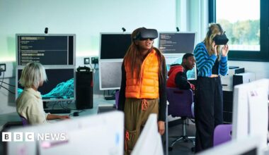 Students in a classroom with computers. Two female students are wearing VR headsets. One student wears an orange gilet and the other a blue stripy cardigan. Behind them are students looking at them and the computers, which show code on the screen.