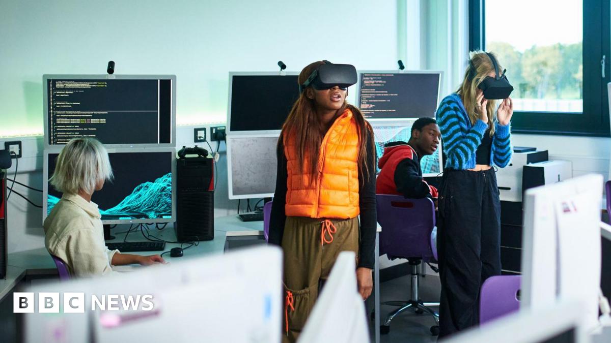Students in a classroom with computers. Two female students are wearing VR headsets. One student wears an orange gilet and the other a blue stripy cardigan. Behind them are students looking at them and the computers, which show code on the screen.