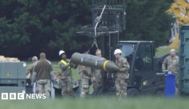 Image of an air base, with a forklift truck in the background.  In the foreground can be seen a number of military personnel in khaki uniforms who are guiding explosive ordnance into position.
