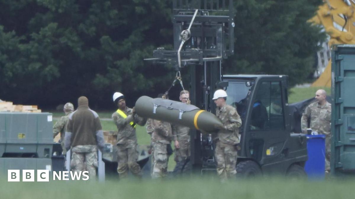 Image of an air base, with a forklift truck in the background.  In the foreground can be seen a number of military personnel in khaki uniforms who are guiding explosive ordnance into position.