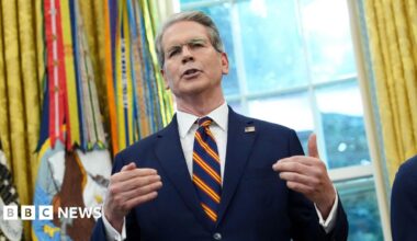 Scott Bessent in the US Whitehouse's Oval Office. He wears a navy suit with a white shirt and striped tie, with the US official ensemble in the background.