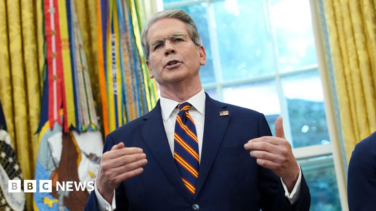 Scott Bessent in the US Whitehouse's Oval Office. He wears a navy suit with a white shirt and striped tie, with the US official ensemble in the background.