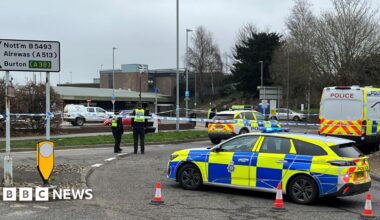 Police cars and a police van and cordon outside the station. At the front of the picture, a police car with blue and yellow markings is positioned and there are traffic cones on the ground. Four police officers are also in the picture.
