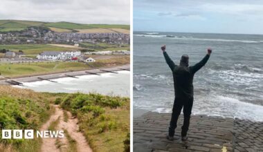 St Bees beach is pictured in the background with groups of houses near the water's edge with groynes lining the beach itself, and in the foreground is a coastal path surrounded by vegetation, on a cloudy day.
