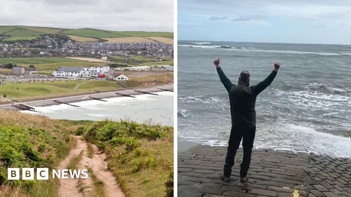 St Bees beach is pictured in the background with groups of houses near the water's edge with groynes lining the beach itself, and in the foreground is a coastal path surrounded by vegetation, on a cloudy day.
