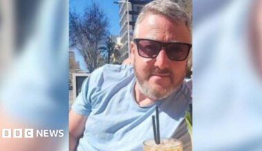 Photo of David Rogers sat outside wearing sunglasses with a drink in front of him. He is wearing a blue t-shirt and has short grey hair and beard.