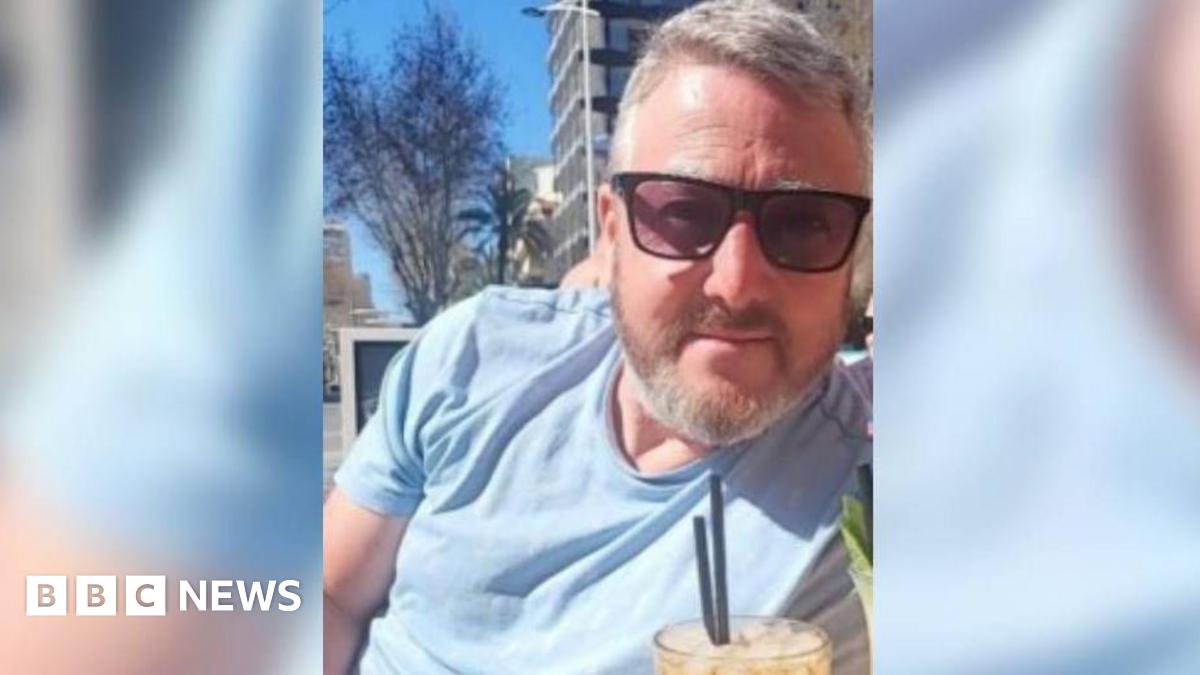 Photo of David Rogers sat outside wearing sunglasses with a drink in front of him. He is wearing a blue t-shirt and has short grey hair and beard.