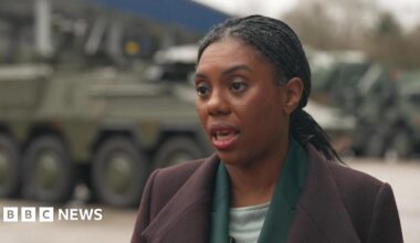 Kemi Badenoch wearing a purple coat, standing in front of a tank.