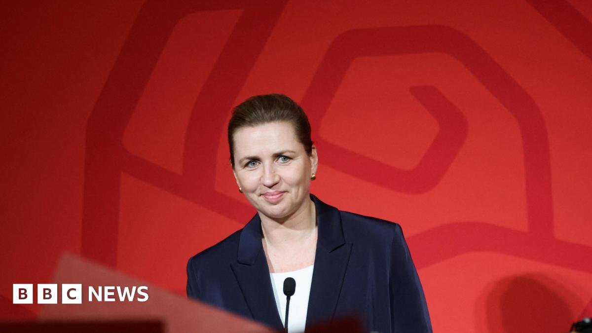 Mette Frederiksen, Denmark's Prime Minister, stands at a podium in front of a red background. She wears a blazer and is smiling slightly.