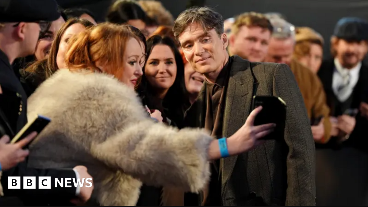 Cillian Murphy, wearing a dark-coloured suit and coat, is posing for a selfie with a woman wearing a fluffy coat. she is leaning over a barrier, and there are a large number of people alongside and behind her.
