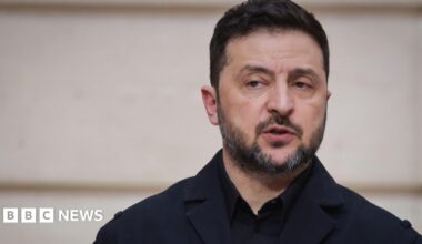 A woman with dark hair and a multiple colour jumper siting on a green sofa inside her living room.