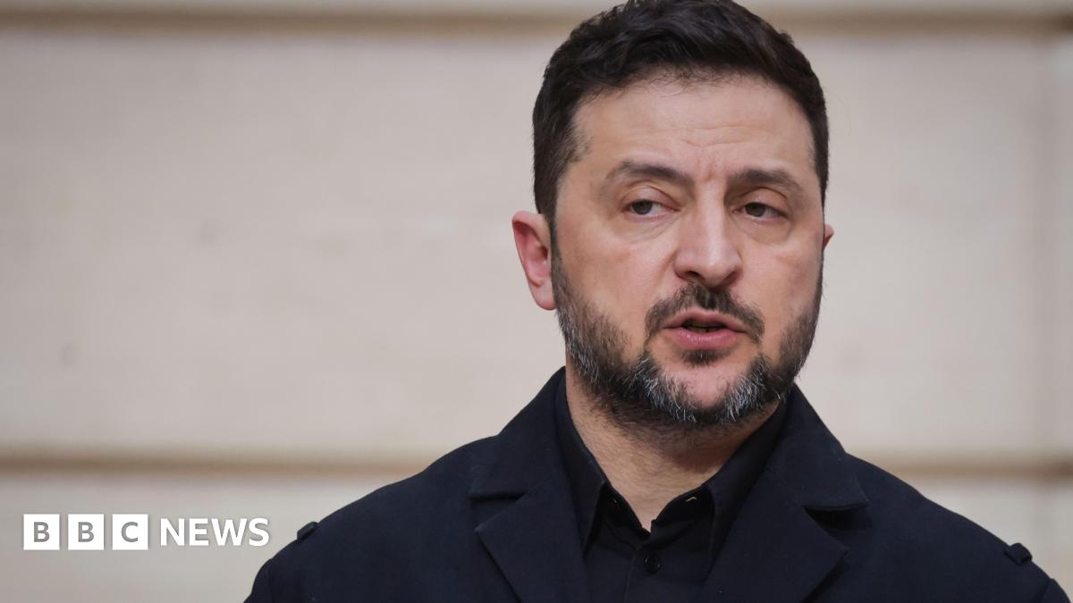 A woman with dark hair and a multiple colour jumper siting on a green sofa inside her living room.