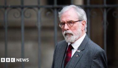 A man with white hair and beard wearing glasses. He is walking past iron gates outside a building. He is wearing a grey suit, white shirt and red tie.