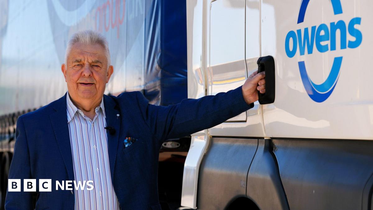 Huw Owen, wearing a blue blazer and striped shirt, stands next to the cab of one of his branded Owens Group lorries. He is holding the door handle and smiling at the camera on a sunny day.