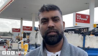 A man with short black and grey hair standing on a petrol forecourt. He is wearing a white T shirt and grey coat and looking down the camera.