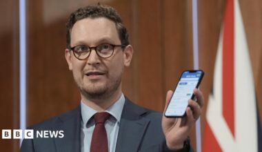 Darren Jones, in dark rimmed glasses and a blue suit with red tie, holds up a smartphone at a Downing Street press conference