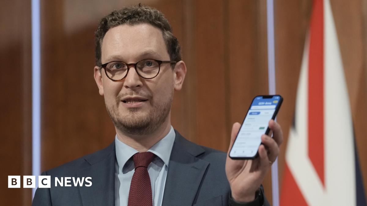 Darren Jones, in dark rimmed glasses and a blue suit with red tie, holds up a smartphone at a Downing Street press conference