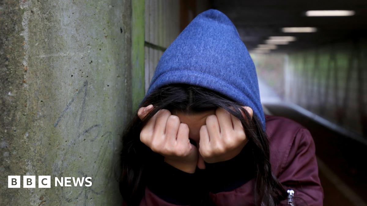 A young women holds her head in her hands, covering her face. She is wearing a blue hoodie and standing in a dark underpass.