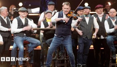 Mike McGrother performs on the violin on stage backed by his choir who are standing behind him with their traditional uniform of waistcoat, white shirts and flat caps.